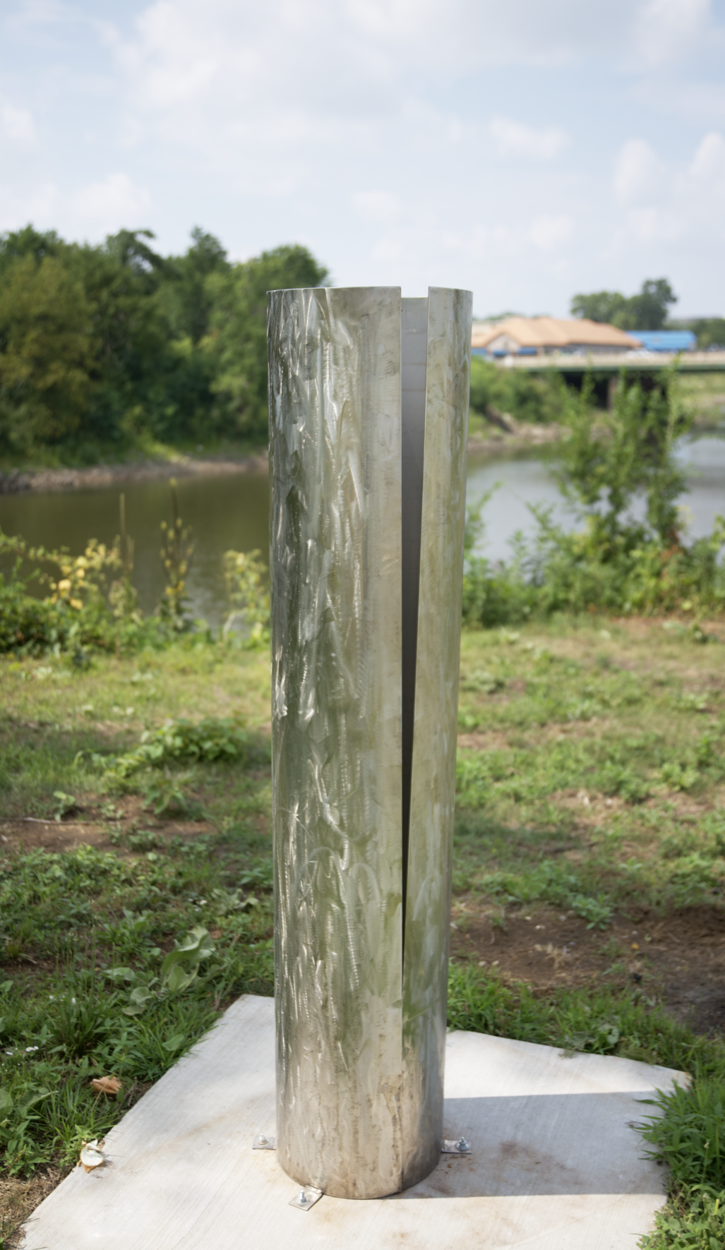 Tall, slender stainless steel sculpture is shown in a park.